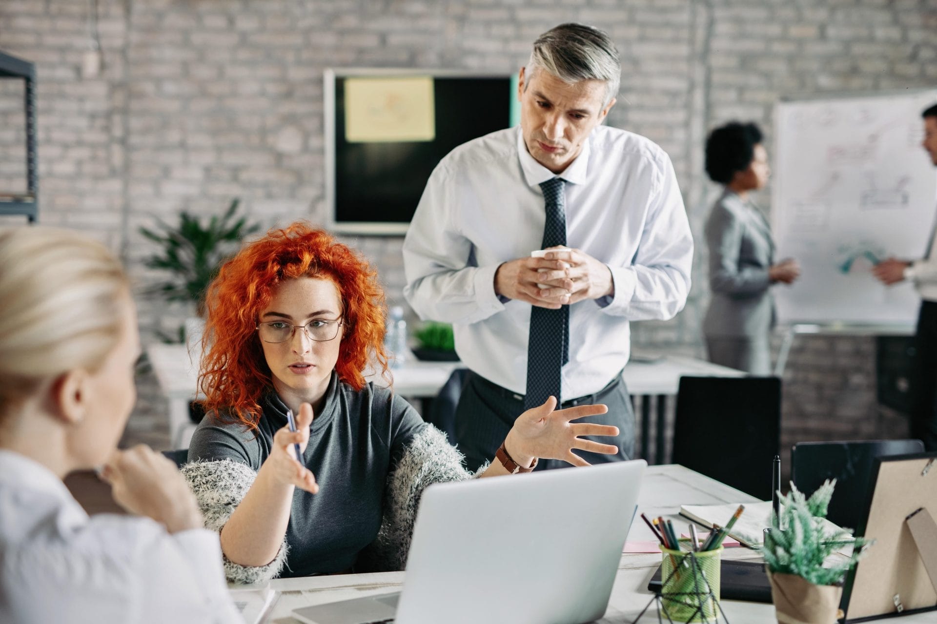 Team members discussing ideas around a laptop during an office meeting, with colleagues presenting at a whiteboard in the background