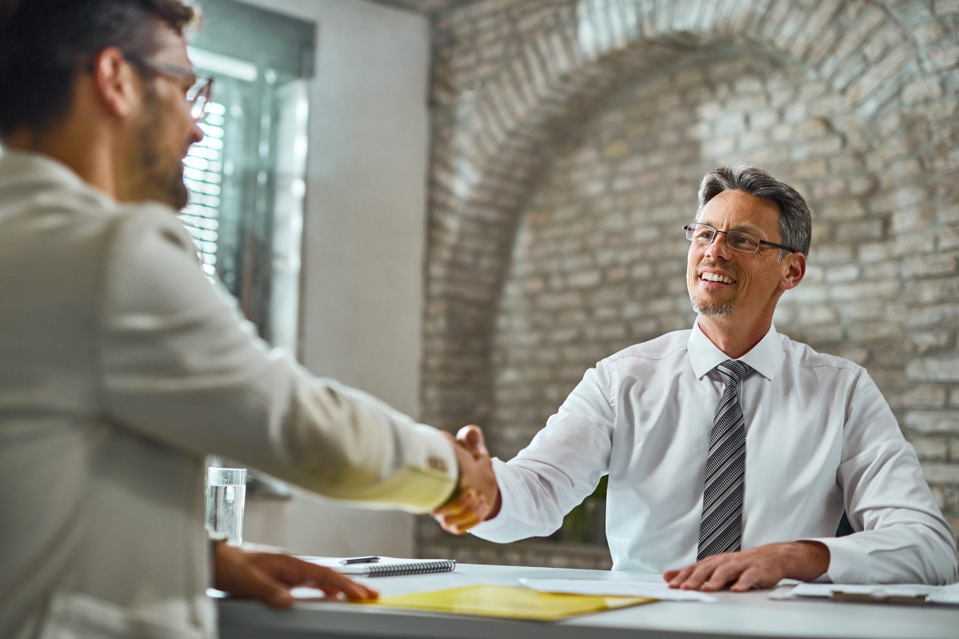 Two businessmen shaking hands across a desk in an office with an exposed brick archway