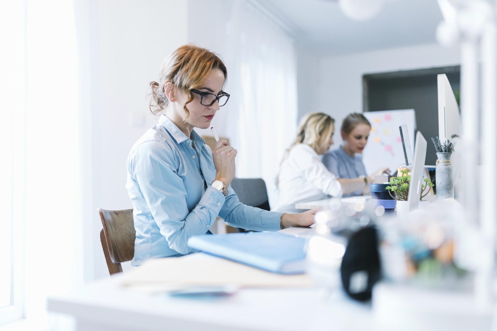 Businesswoman in glasses thinking at her desk while colleagues collaborate at a whiteboard in the background